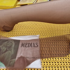 Puede incluir: Primer plano de una media de nylon marrón con una etiqueta que dice "MEDIAS INDE MALLABLES". La media está sobre una superficie con un patrón amarillo y marrón.
