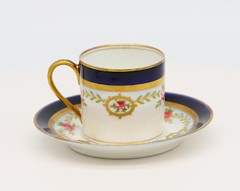 Może przedstawiać: Biały porcelanowy kubek i spodek z niebieską i złotą obwódką i kwiatowym wzorem. Kubek ma złoty uchwyt.