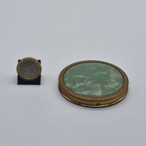 Puede incluir: Un compacto ovalado vintage con un marco de metal dorado y una superficie verde claro veteada. El compacto está junto a una moneda de un euro sobre un soporte negro. El compacto mide aproximadamente 7,6 cm por 5 cm.