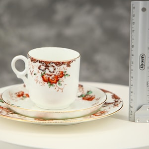 Puede incluir: Un juego de taza y platillo de porcelana blanca con un diseño floral de flores naranjas, hojas verdes y cintas marrones, con bordes dorados. Una regla de plástico gris colocada al lado muestra una altura de 7,5 cm.