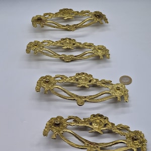 Puede incluir: Cuatro tiradores de cajones de color dorado adornados con un diseño floral. Cada tirador tiene una forma curva y detalles intrincados.