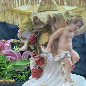 May include: A detailed close-up of a porcelain lamp base featuring a cherub figure, red raspberries, and green leaves. The base has a cream-coloured, ruffled design with gold accents. The lamp shade is visible in the background.