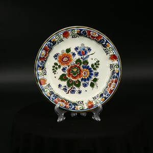 Può includere: Un piatto decorativo in ceramica con base bianca, caratterizzato da un motivo floreale arancione, blu e verde. Il bordo è decorato con un motivo blu e rosso. Il piatto è esposto su un supporto trasparente.