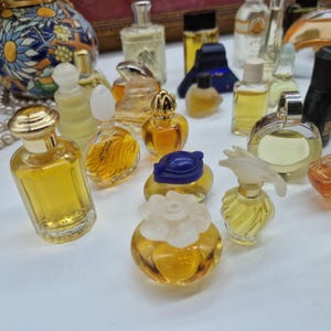 K&ouml;nnte beinhalten: Eine Sammlung von Miniatur-Parf&uuml;mflaschen in verschiedenen Formen und Gr&ouml;&szlig;en. Die Flaschen sind mit einer goldenen Fl&uuml;ssigkeit gef&uuml;llt und haben dekorative Verschl&uuml;sse in Gold, Blau und Wei&szlig;. Die Flaschen sind auf einer wei&szlig;en Oberfl&auml;che angeordnet.