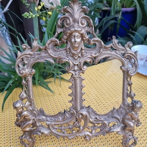 May include: Ornate brass-colored picture frame with a central female face and cherub figures. The frame has an elaborate, scrolling design with a detailed, antique aesthetic. The frame is designed to stand upright.
