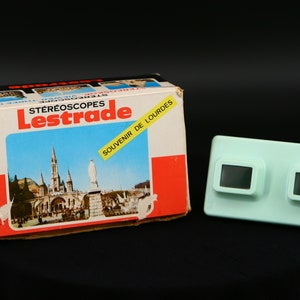 Puede incluir: Una caja azul y blanca con el texto "Stereoscopes Lestrade Souvenir de Lourdes" en la parte delantera. La caja contiene un visor estereoscópico vintage con dos agujeros de visión negros.