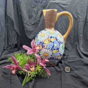 Puede incluir: Jarra de cerámica decorativa con un diseño floral con margaritas blancas y campanillas azules sobre un fondo azul. La jarra tiene una parte superior marrón y un asa dorada. Orquídeas rosas y vegetación frente a la jarra.