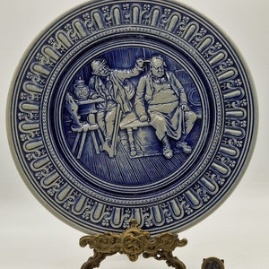 Può includere: Un piatto in ceramica blu e bianco con un design in rilievo di due uomini seduti a un tavolo. Il piatto ha un bordo decorativo con un motivo ripetitivo. Il piatto è su un supporto in metallo dorato.