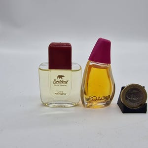 Può includere: Due flaconi di profumo vintage. Uno è un flacone quadrato con un tappo rosso scuro, etichettato "Rockford Eau de Toilette 6ml Atkinsons". L'altro è un flacone a forma di goccia con un tappo magenta, etichettato "Poivre". Una moneta poggia su un supporto nero.