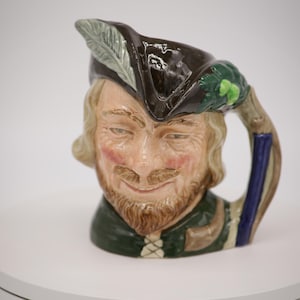 Puede incluir: Una taza de cerámica con un personaje con un sombrero marrón, hojas verdes y un asa azul y blanca. El personaje tiene barba y bigote marrones y lleva una chaqueta verde y marrón.