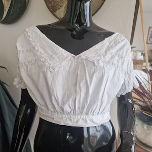 May include: A white, vintage crop top with a V-neck and short sleeves. The top has delicate lace trim along the neckline and sleeves. The fabric appears to be a lightweight cotton or linen, gathered at the waist.