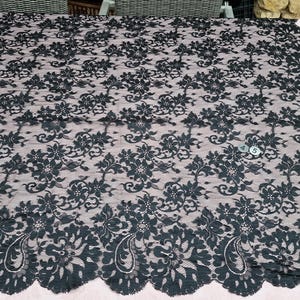 May include: Black lace fabric with a floral pattern. The lace has a scalloped edge and is laid out on a light pink surface. The intricate design features flowers and leaves, creating a delicate and elegant look. This fabric is suitable for various sewing projects.