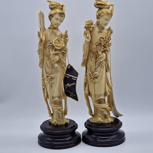 Puede incluir: Dos estatuas de color marfil de mujeres con vestimenta tradicional, cada una sosteniendo flores. Se alzan sobre bases de madera oscura. Una estatua tiene una etiqueta que dice "Marco's". Las estatuas tienen rasgos faciales detallados y vestiduras fluidas.