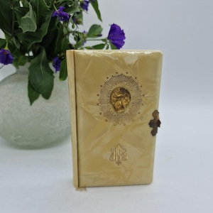 Könnte beinhalten: Cremefarbenes, rechteckiges Buch mit einem dekorativen goldenen Totenkopf-Emblem und Verschluss. Das Cover hat ein Rautenmuster. Eine kleine Quaste hängt von unten. Das Buch ist wahrscheinlich ein Tagebuch oder ein Notizbuch.