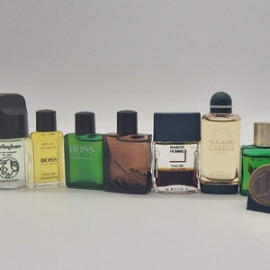 Puede incluir: Una colección de frascos de colonia en miniatura de varios colores y diseños. Los frascos están dispuestos en una fila sobre una superficie blanca. Algunos de los frascos tienen etiquetas con los nombres de las fragancias, como "Boss", "Barbour Homme" y "Freelude".