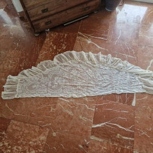 May include: Cream lace valance with a scalloped edge and floral design. The sheer fabric valance is displayed on a red marble floor. This window treatment accessory features a delicate, openwork pattern, suitable for a bedroom or living room.