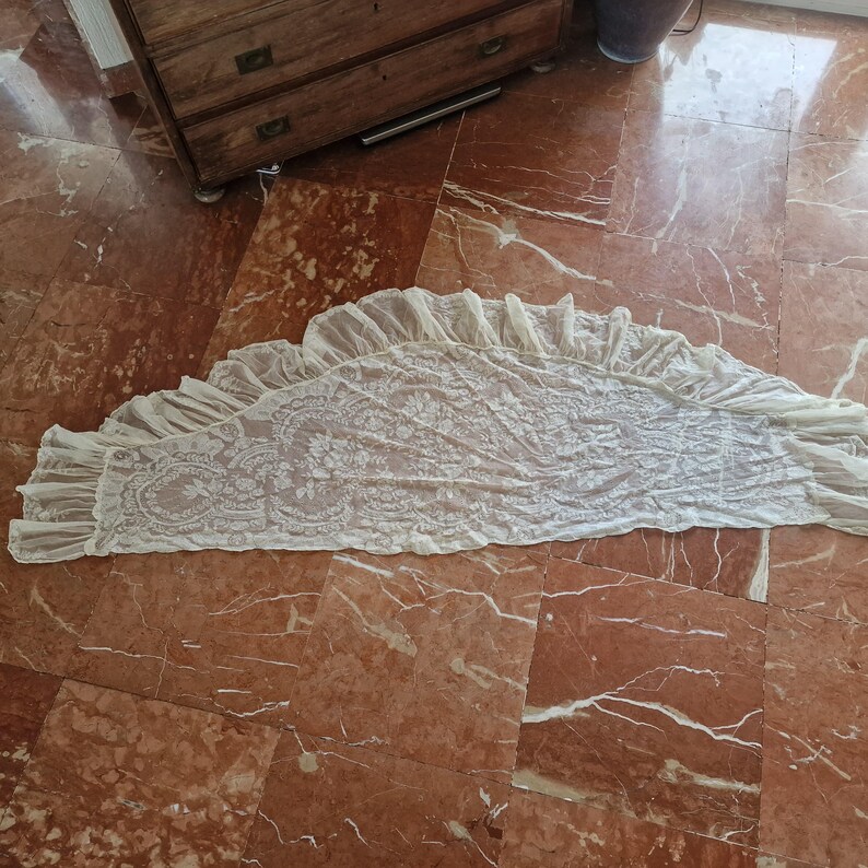 May include: Cream lace valance with a scalloped edge and floral design. The sheer fabric valance is displayed on a red marble floor. This window treatment accessory features a delicate, openwork pattern, suitable for a bedroom or living room.