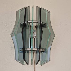 Puede incluir: Aplique de pared vintage con un diseño geométrico. La lámpara presenta múltiples paneles de vidrio transparente y tintado, sujetos por detalles de metal cromado. La lámpara está montada en una pared y tiene un cable de alimentación.