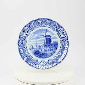 Puede incluir: Un plato decorativo azul y blanco con una escena de molino de viento. El plato tiene un borde festoneado y un diseño de borde detallado. El molino de viento es el punto focal, con otros edificios y un cuerpo de agua en el fondo.