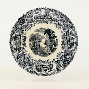 May include: A white ceramic plate with a black and white design. The design features a landscape scene with a building, trees, and water. The plate has a decorative border.