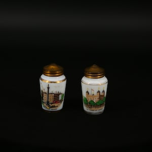 Op de afbeelding: Twee kleine zout- en peperstrooiers van wit keramiek met goudkleurige deksels en randen. Elke strooier heeft een kleurrijke illustratie van een Londense bezienswaardigheid. Eén strooier toont Trafalgar Square, de andere de Tower of London.