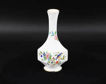 Puede incluir: Un jarrón de porcelana blanca con un borde dorado y un diseño floral con pájaros y flores. El jarrón tiene una forma geométrica con un cuello estrecho y una base ancha.