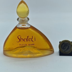 Pode incluir: Um frasco de perfume vintage com uma tampa amarela em forma de gota. A garrafa é preenchida com um líquido dourado e tem a palavra "Shafali" em escrita vermelha. As palavras "FLEUR RARE" estão abaixo. Há uma moeda ao lado.