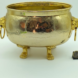 Puede incluir: Un macetero de metal dorado con asas de cabeza de león y tres patas. El macetero tiene un acabado martillado y es de forma ovalada.