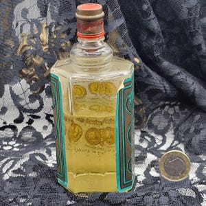 May include: A vintage perfume bottle with a clear glass body and a red and brown cap. The bottle has a rectangular shape with teal and brown labels featuring gold coin illustrations. The liquid inside is yellow. A coin is next to the bottle.