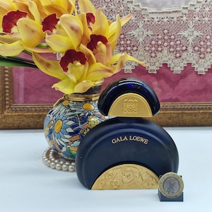 May include: A navy blue perfume bottle with gold detailing, labelled "GALA LOEWE". Beside it, a small vase holds yellow and red flowers. A coin is positioned in front of the bottle.