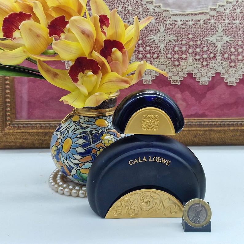 May include: A navy blue perfume bottle with gold detailing, labelled "GALA LOEWE". Beside it, a small vase holds yellow and red flowers. A coin is positioned in front of the bottle.