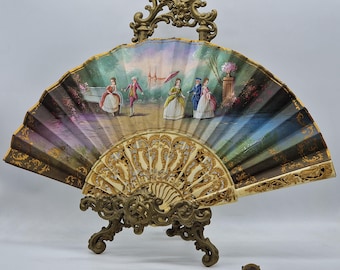 May include: An antique hand fan with a painted scene of figures in period clothing, displayed on a decorative brass stand. The fan features a cream-colored, intricately carved base and a painted upper section with gold accents. The scene depicts a park setting.