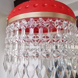 Puede incluir: Una lámpara de araña roja y dorada con gotas de cristal transparentes. La lámpara de araña tiene una parte superior redonda roja con ribete dorado y una serie de niveles de plástico transparente con un patrón de estrella.