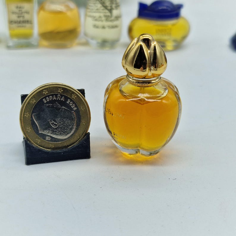 K&ouml;nnte beinhalten: Ein kleiner, klarer Parf&uuml;mflakon mit goldfarbenem Verschluss und gelber Fl&uuml;ssigkeit. Eine M&uuml;nze mit der Aufschrift "ESPA&Ntilde;A 2024" befindet sich neben der Flasche. Weitere Parf&uuml;mflakons sind im Hintergrund zu sehen.