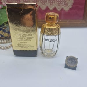 Könnte beinhalten: Ein kleiner, klarer Glasparfümflakon mit goldenem Verschluss und dem Wort "CHAMPAGNE" auf der Vorderseite. Die Flasche steht neben einer goldfarbenen Schachtel mit Text und einer Münze.