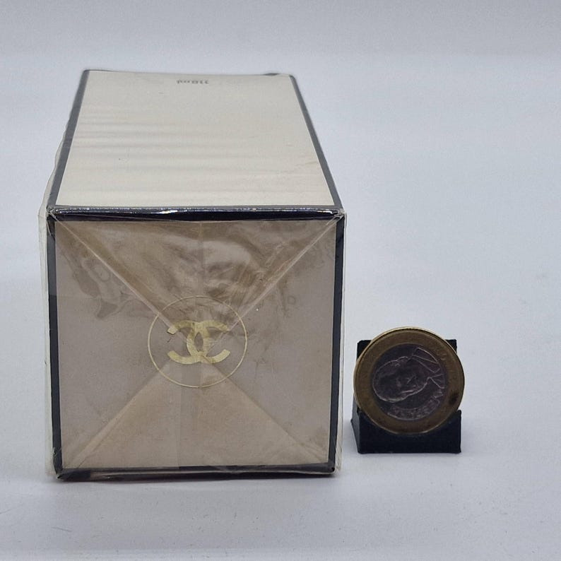 May include: A rectangular perfume box wrapped in clear plastic. The box is beige with a gold logo and text. A coin is next to the box for size comparison.