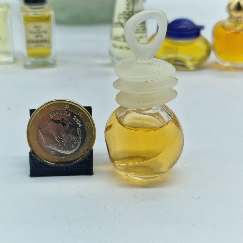K&ouml;nnte beinhalten: Kleine, transparente Parf&uuml;mflasche mit hellbrauner Fl&uuml;ssigkeit, gekr&ouml;nt von einem Design aus mattierten, gestapelten Scheiben und einer Schlaufe. Eine M&uuml;nze befindet sich neben der Flasche. Weitere Parf&uuml;mflaschen sind im Hintergrund.