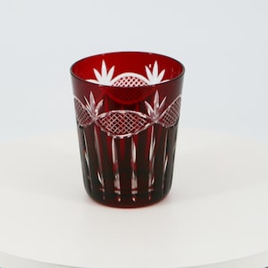 Può includere: Un bicchiere da whisky in vetro rosso scuro con un design in cristallo tagliato. Il bicchiere ha un motivo chiaro e intricato inciso sulla superficie.