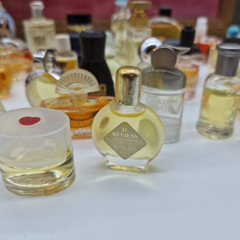 K&ouml;nnte beinhalten: Eine Sammlung von Vintage-Parf&uuml;mflaschen in verschiedenen Formen und Gr&ouml;&szlig;en. Die Flaschen sind mit einer gelben Fl&uuml;ssigkeit gef&uuml;llt. Eine Flasche hat einen goldenen Verschluss und ein Etikett mit der Aufschrift "Je Reviens Worth".