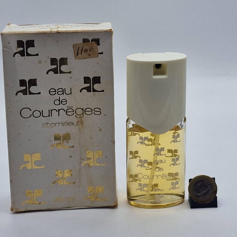 May include: A vintage Eau de Courr&egrave;ges atomizer perfume bottle with its original box. The box is off-white with gold-colored logos and text. The perfume bottle is clear with a cream-colored top and contains a yellow liquid. A coin is next to the bottle.