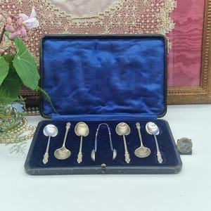 Puede incluir: Un juego de seis cucharas de plata y unas pinzas de azúcar, presentado en un estuche negro vintage con forro de terciopelo azul. Las cucharas tienen mangos ornamentados y cuencos redondeados. El estuche está abierto.