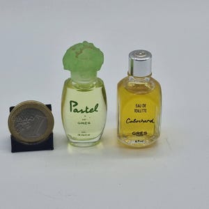 Puede incluir: Dos frascos de perfume en miniatura, uno con la etiqueta "Pastel de Grès" con un tapón verde claro, y el otro "Cabochard Eau de Toilette". Una moneda de euro está al lado de los frascos. Los frascos están llenos de un líquido amarillo.