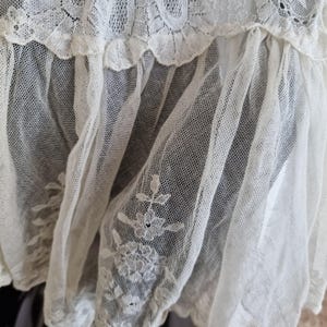 May include: Close-up of a delicate, cream-coloured lace fabric. The lace features a floral embroidered design and a scalloped edge. The fabric is layered, creating depth and texture. The overall aesthetic is vintage and romantic.