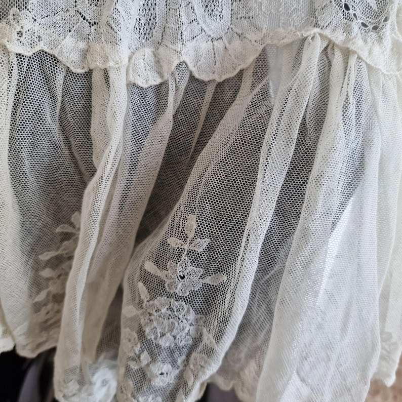 May include: Close-up of a delicate, cream-coloured lace fabric. The lace features a floral embroidered design and a scalloped edge. The fabric is layered, creating depth and texture. The overall aesthetic is vintage and romantic.