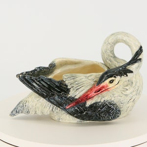 May include: Ceramic stork planter in a resting position. The bird is primarily white with black wing accents and a red beak. The planter has a shallow, open bowl for holding plants or other items.