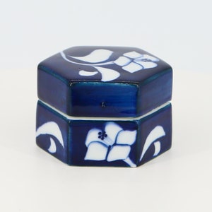 Pode incluir: Uma pequena caixa de cerâmica octogonal azul e branca com um design floral. A caixa tem uma tampa e é decorada com flores e folhas brancas sobre um fundo azul escuro.