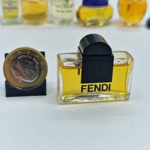 K&ouml;nnte beinhalten: Ein Miniatur-Fendi-Parf&uuml;mflakon mit schwarzem Verschluss und wei&szlig;em Markennamen. Die Flasche enth&auml;lt eine gelbe Fl&uuml;ssigkeit. Neben der Flasche liegt eine M&uuml;nze.