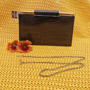 Può includere: Una pochette vintage marrone con una cornice color oro e una tracolla a catena staccabile. La pochette ha una trama simile a quella del coccodrillo e una forma rettangolare. L'etichetta recita "Cocodrilo Rosa". Due fiori arancioni e rossi sono accanto alla pochette.