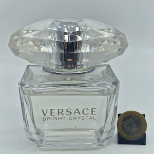 Puede incluir: Un frasco de perfume Versace Bright Crystal de vidrio transparente. El frasco tiene un tapón facetado en forma de diamante y un cuerpo rectangular. El frasco de perfume está junto a una moneda.