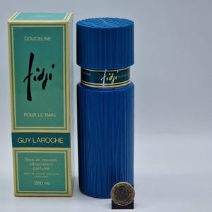 Puede incluir: Una botella y caja de perfume Fiji de Guy Laroche azules. La botella tiene un diseño texturizado y forma cilíndrica. La caja es verde azulado con el nombre "Fiji" en una fuente estilizada. La caja también dice "Pour le Bain" y contiene 260 ml.
